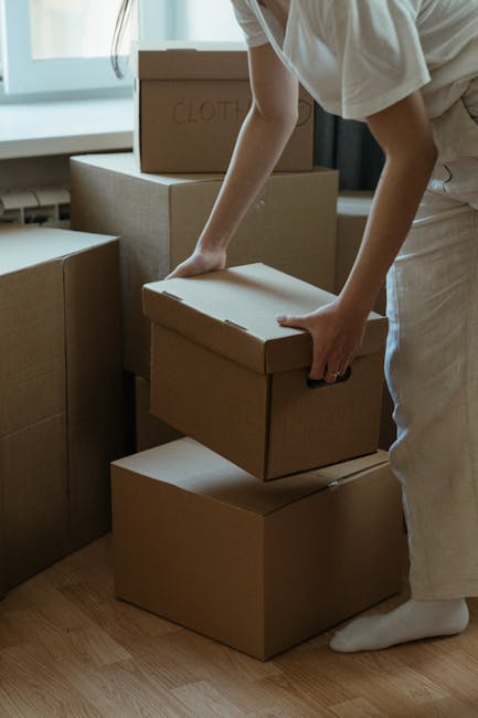 A person, partially visible and dressed in neutral-colored clothing, is shown in an indoor setting arranging and stacking medium-sized cardboard boxes on top of each other. One box in their hands has the word 'CLOTH' handwritten on it, indicating its contents, while other boxes are placed on a wooden floor near a window, suggesting a packing process during a house relocation. The scene depicts an organized packing area inside a home, with natural light illuminating the space and furniture or other moving supplies not visible in the frame. The use of cardboard boxes and careful stacking reflects the packing and moving preparations typical of a furniture and house removal service, such as that provided by Man and Van Coulsdon.