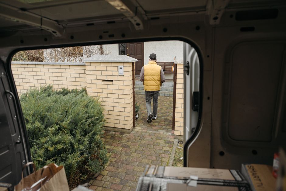 A man wearing a black and grey cap, a black vest over a blue long-sleeve shirt, is inside a white panel van with open rear doors. He is holding a rectangular cardboard box wrapped in brown packing pap