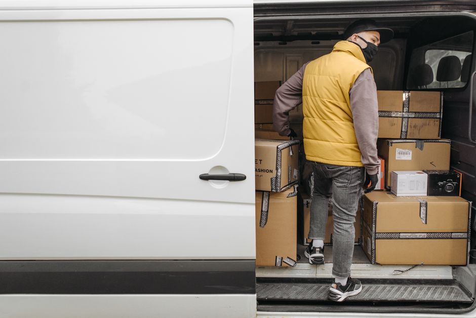 A man wearing a face mask, baseball cap, yellow and grey jacket, and grey trousers stands on a brick driveway next to a white commercial van parked outside a residential property. The van is positione